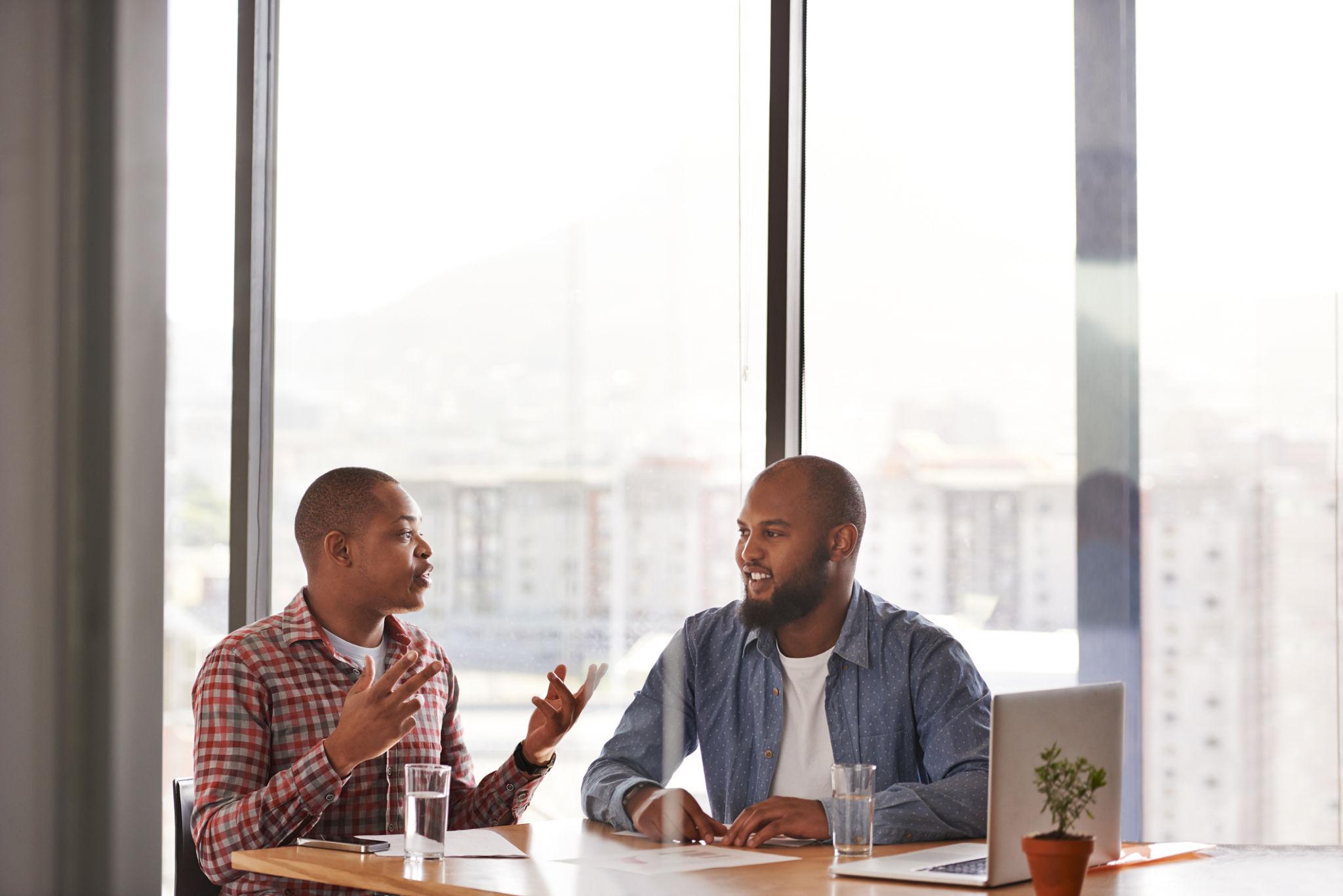 Two colleagues talking at a table beside a tall office window